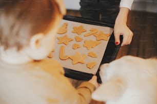 Family baking gingerbread cookies. Cute little daughter, dog and mother holding tray with christmas cookies close up. Family holiday preparation, xmas culinary. Best friends, authentic moment