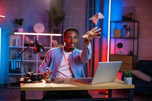 Happy young man in casual wear throwing paper plane while taking break during remote work on laptop. Smiling black guy sitting at desk during evening time and having fun.