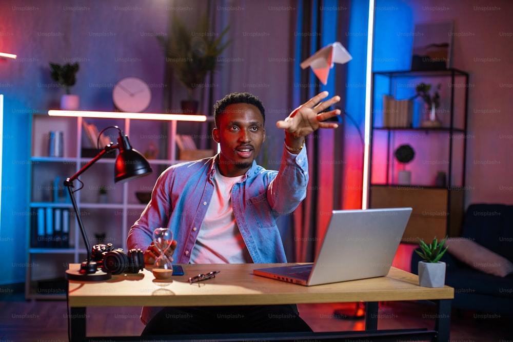 Happy young man in casual wear throwing paper plane while taking break during remote work on laptop. Smiling black guy sitting at desk during evening time and having fun.