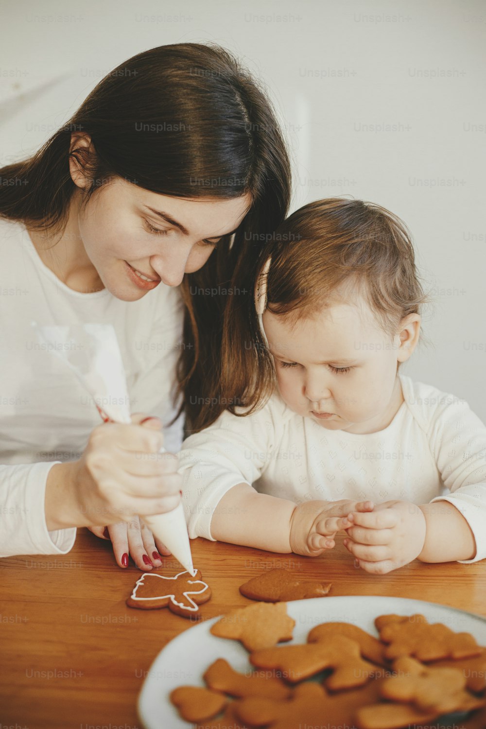 Cute little daughter and mother together decorating christmas gingerbread cookies with icing on wooden table in modern room.  Mommy daughter authentic lovely moments. Holiday preparations