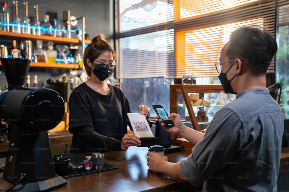 Asiatischer Mann trägt während der COVID-19-Pandemie eine schützende Gesichtsmaske mit Smartphone-Scan-Barcode Machen Sie kontaktloses Bezahlen im Café. Kleinunternehmen mit Mobile-Banking-Konzept für elektronisches Bezahlen