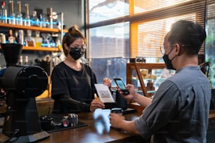 Un homme asiatique porte un masque de protection pendant la pandémie de COVID-19 à l’aide d’un smartphone scannant le code-barres pour effectuer un paiement sans contact dans un café. Petites entreprises avec concept de paiement électronique mobile