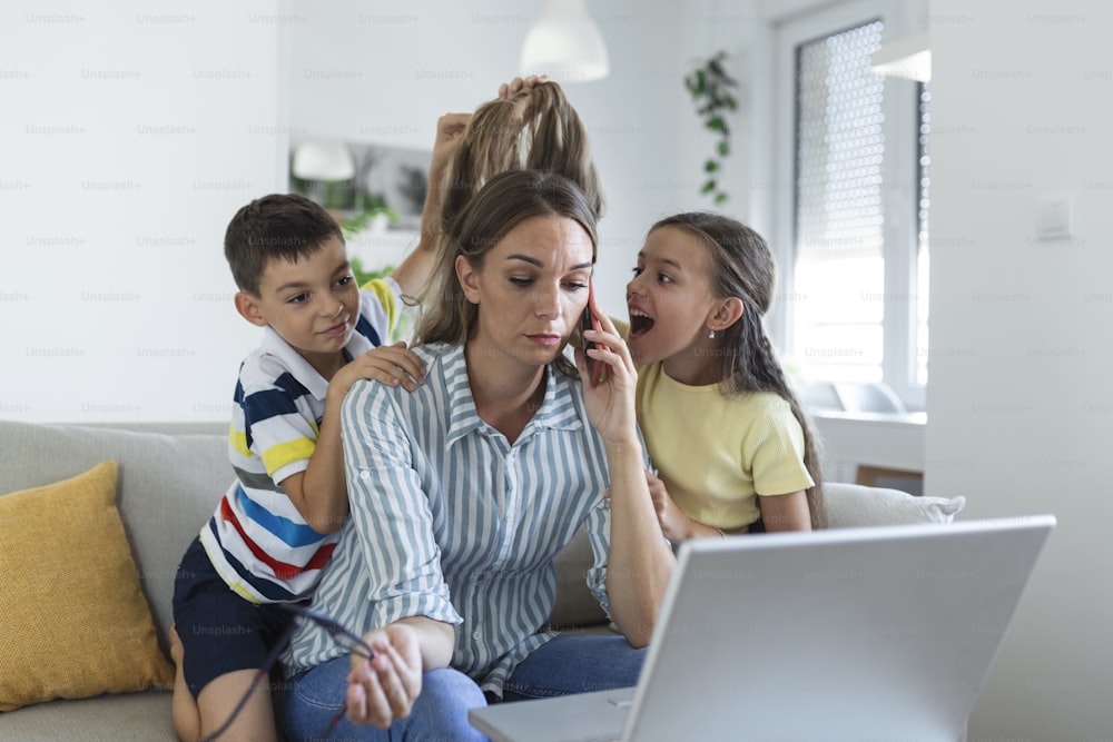 Mulher que trabalha em casa durante a quarentena com seu filho pequeno e filha no sofá e gritando, exigindo atenção