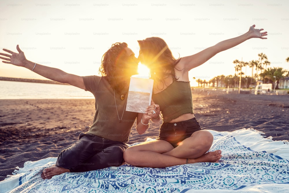Schönes glückliches Paar bei einem Date, küsst sich und zeigt ein Buch am tropischen Strand. Sommerferien-Vibes. Sonnenuntergangszeit.