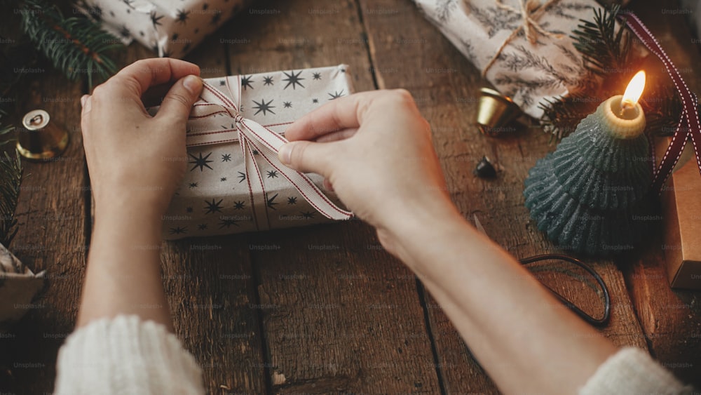 Hands wrapping stylish christmas gift in craft paper on rustic wooden table with candles, scissors, fir branches. Merry Christmas! Stylish scandinavian xmas presents, atmospheric moody image