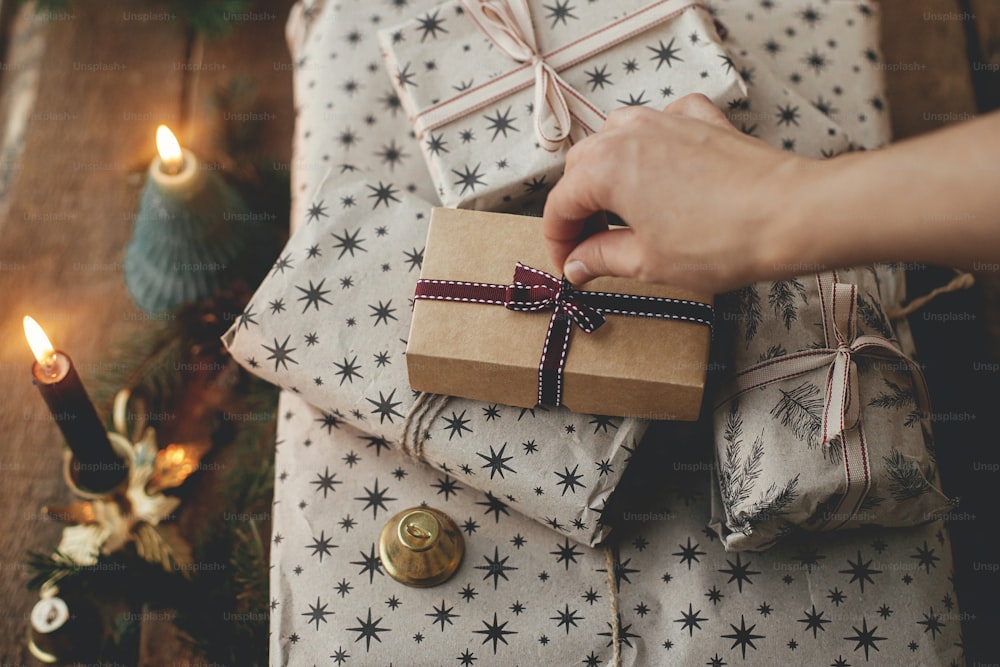 촛불, 종, 전나무 가지가 있는 소박한 나무 테이블에 종이에 싸인 세련된 크리스마스 선물을 손으로 엽니다. 세련 된 스칸디나비아 크리스마스 선물, 분위기 있는 변덕스러운 이미지. 즐거운 성탄절!
