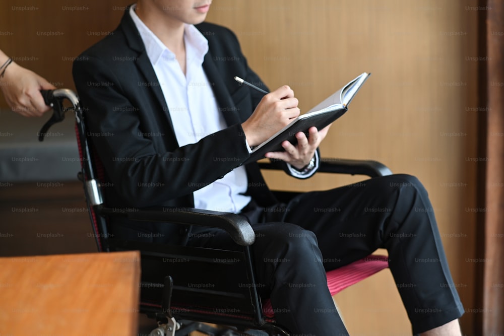 Hombre de negocios discapacitado en silla de ruedas sosteniendo un bolígrafo y escribiendo alguna información en un cuaderno.