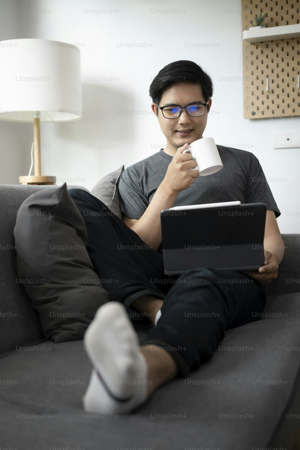 Young Asian man drinking coffee and working with computer tablet on couch.