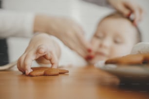 木製のテーブルの上で焼きたてのジンジャーブレッドクッキーをつかむかわいい女の子。本物の素敵な瞬間、休日の準備。愛らしい幼児の女の子がお母さんとクリスマスクッキーを作る