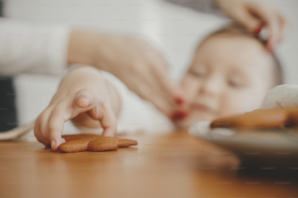Cute little girl grabbing freshly baked gingerbread cookie on wooden table close up. Authentic lovely moment, holiday preparations. Adorable toddler girl making christmas cookies with mother