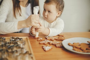 Cute little daughter and mother decorating christmas gingerbread cookies with icing on wooden table, close up. Family time together, xmas holiday preparations. Mommy daughter authentic moment