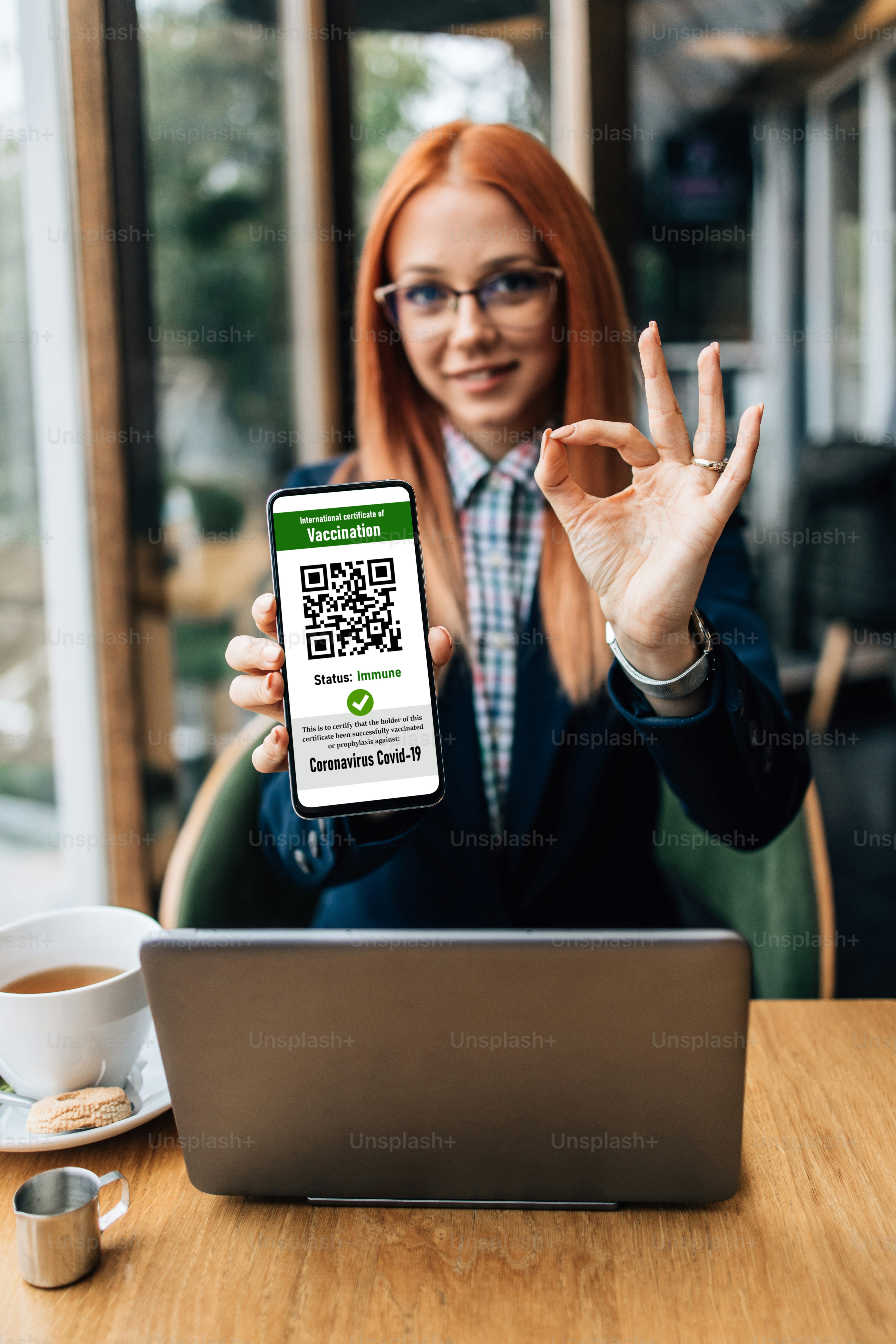 Young business woman or student sitting in bar or restaurant and using laptop. She is also holding smart phone with electronic Covid -19 or Coronavirus vaccination certificate. She is safe and positive and showing OK sign with her hand.