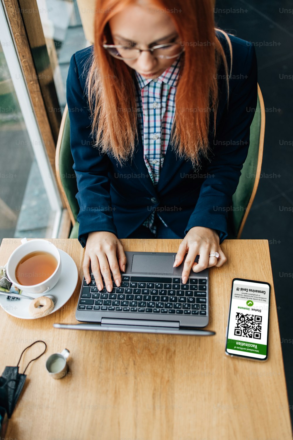 Young attractive business woman or student sitting in a cafe bar or restaurant and using her laptop. Her smart phone is turned on and on screen is visible Covid-19 or Coronavirus vaccination electronic certificate that can be scanned by a waiter.