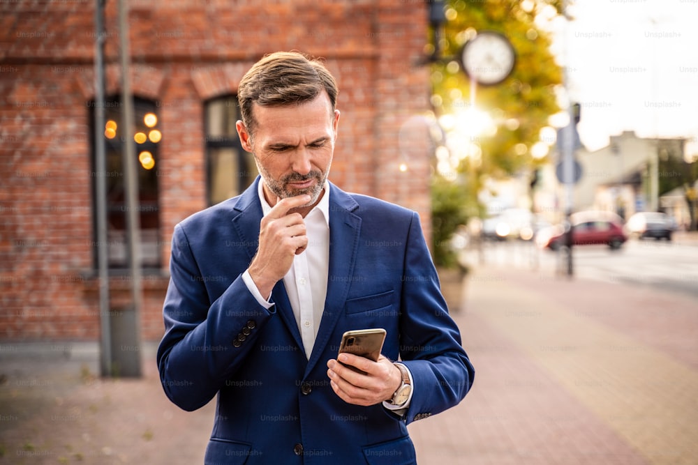 Photo of handsome businessman using mobile phone outdoor on the city street. Thoughtful businessman in elegant suit working on smartphone.