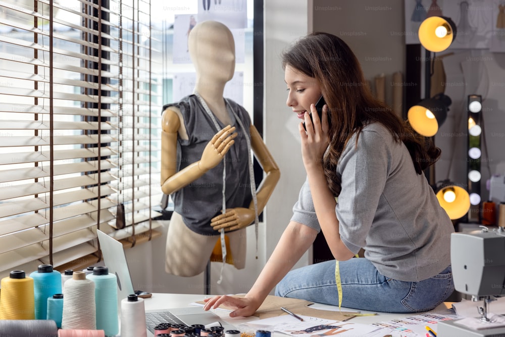 Millennial woman dressmaker making a phone call to her partner while holding her smartphone to her ear. A designer decorator speaks with a client about model features and color choices.
