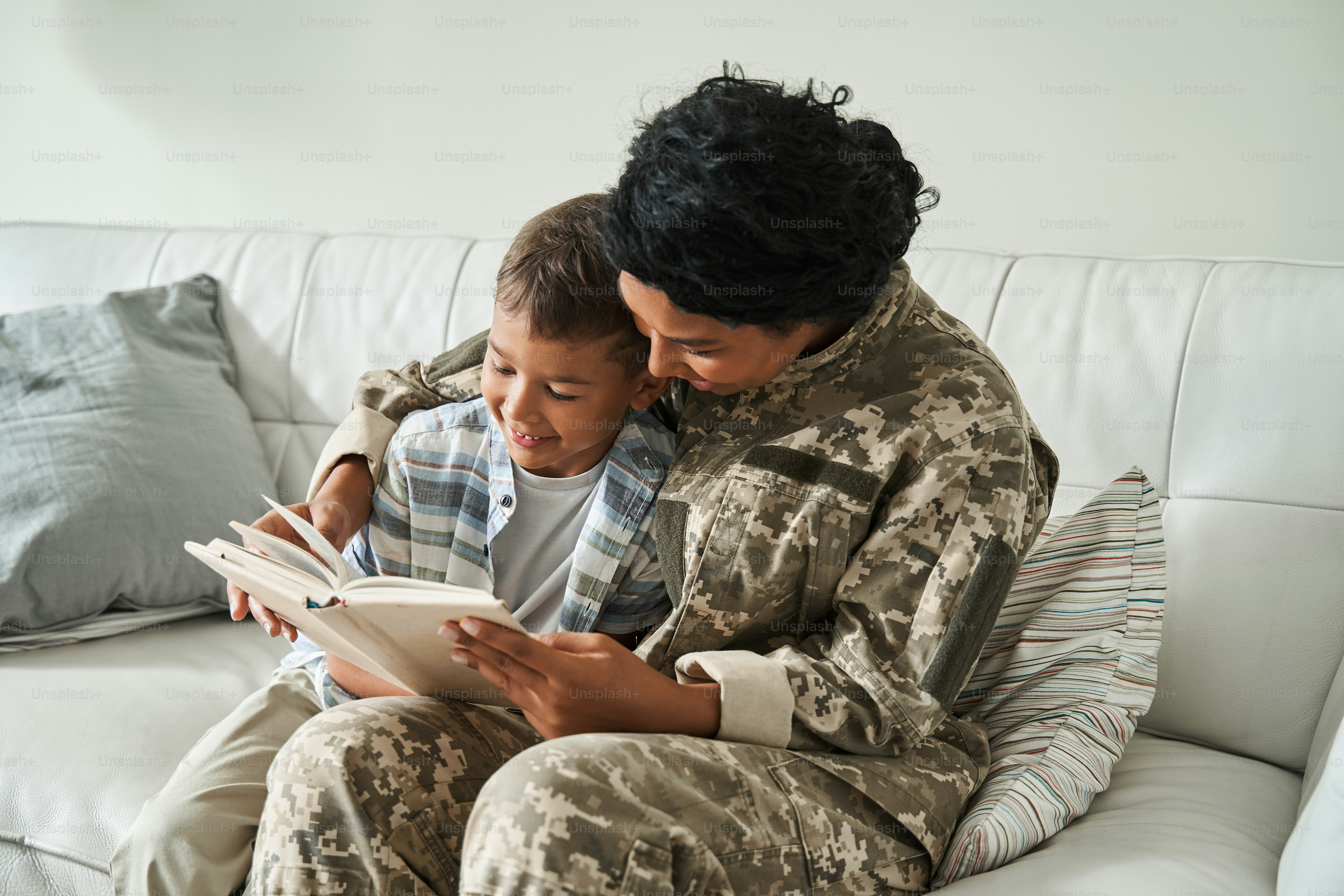 Garoto animado animado para a chegada de suas mães. Mulher multirracial vestindo uniforme lendo livro enquanto sentada no sofá. Conceito de relações familiares