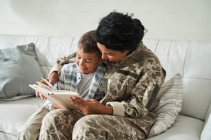 Lively kid excited for his moms arrival. Multiracial woman wearing uniform reading book while sitting at the sofa. Family relationships concept