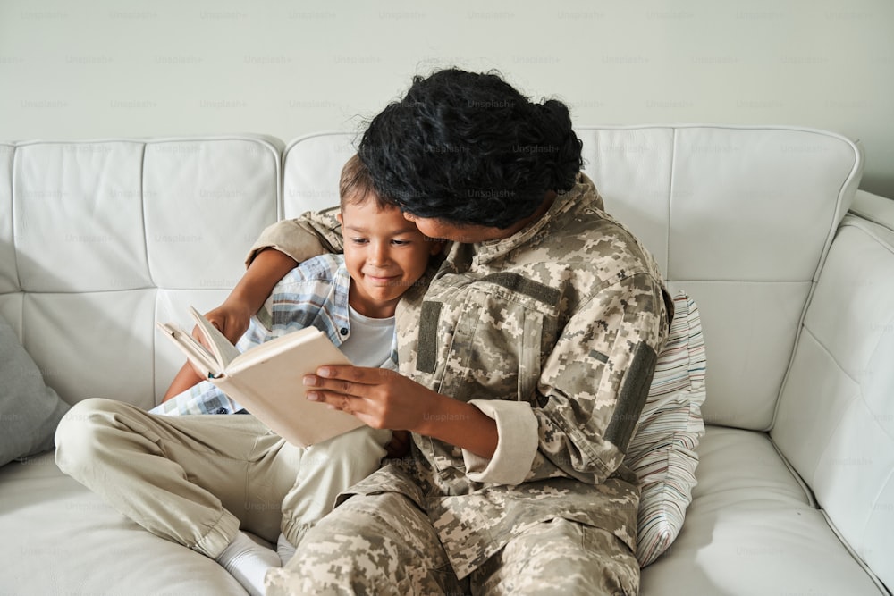 Millennial female soldier returning home to her son. Boy embracing with his mother and reading book together. Family reunion concept
