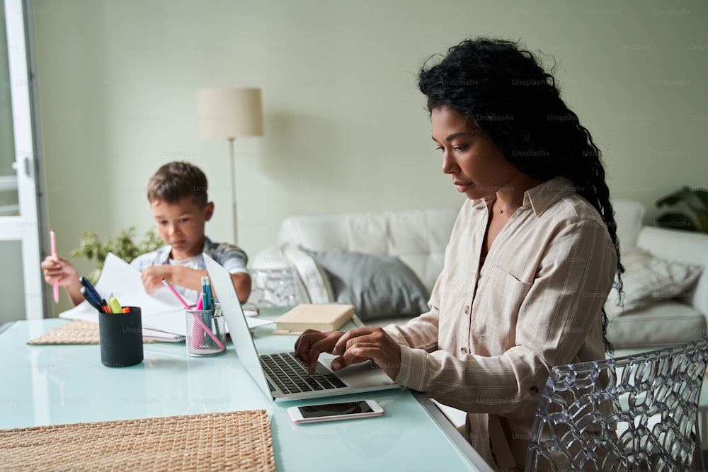 Black mother using laptop computer and her son drawing with pencils at table. Modern successful business woman. Concept of remote work. Family relationships. Interior of living room at modern flat