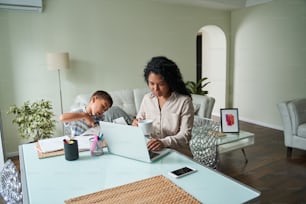 Madre negra usando una computadora portátil y su hijo cortando papel con tijeras en la mesa. Mujer de negocios moderna bebiendo té o café de la taza. Concepto de trabajo remoto. Relaciones familiares. Piso moderno