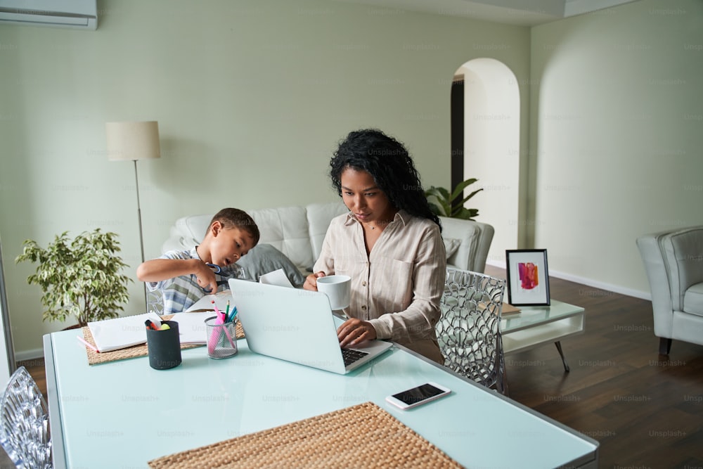 Black mother using laptop computer and her son cutting paper with scissors at table. Modern business woman drinking tea or coffee from cup. Concept of remote work. Family relationships. Modern flat