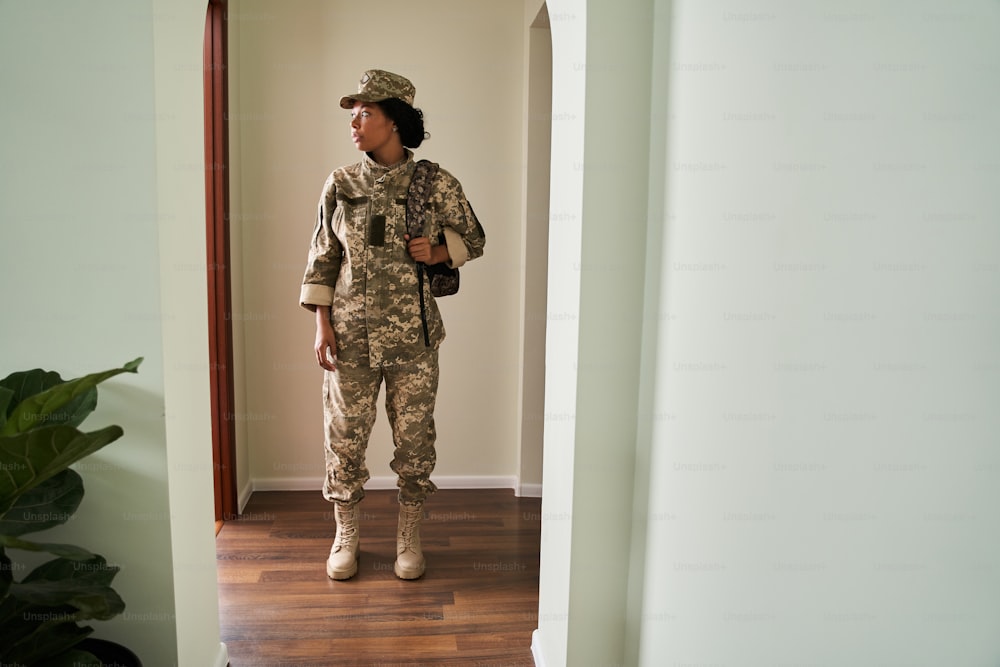 To the war. Full length view of the soldier woman wearing military uniform holding backpack and looking at the door with sad emotions while going to fight