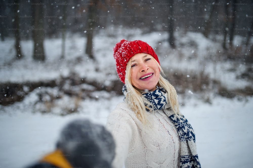 Headshot portrait of happy senior woman with hat and outdoors standing in snowy nature, looking at camera.