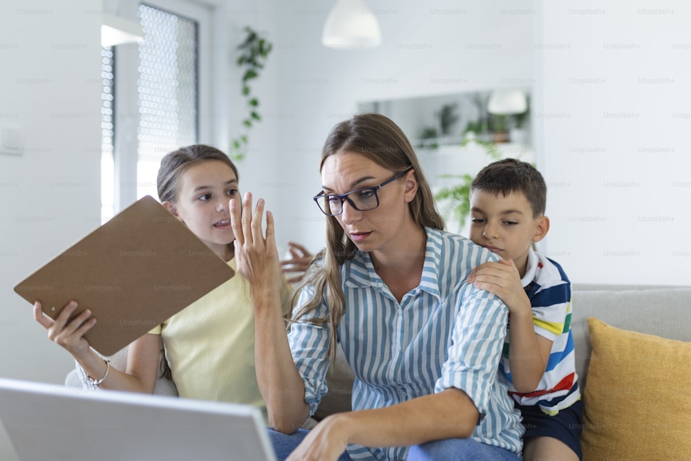 Frau, die während der Quarantäne von zu Hause aus mit ihrem kleinen Sohn und ihrer Tochter auf der Couch arbeitet und schreit und Aufmerksamkeit fordert