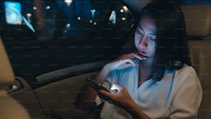 패션 사무복을 입은 성공적인 젊은 아시아 사업가는 밤에 도시 현대 도시의 뒷좌석에 앉아 스마트폰을 사용하여 늦게까지 일하고 있다. 사람들 직업 소진 증후군 개념.