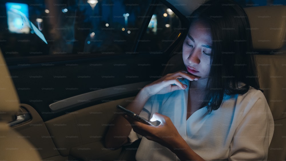 Giovane imprenditrice asiatica di successo in abiti da ufficio di moda che lavora fino a tardi usando lo smartphone sul sedile posteriore dell'auto nella città moderna urbana di notte. Concetto di sindrome da burnout professionale delle persone.