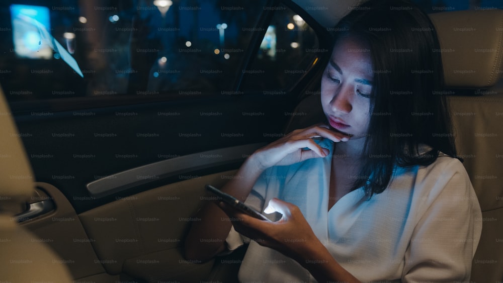 Successful young Asia businesswoman in fashion office clothes working late using smart phone in sitting back seat of car in urban modern city in night. People occupational burnout syndrome concept.