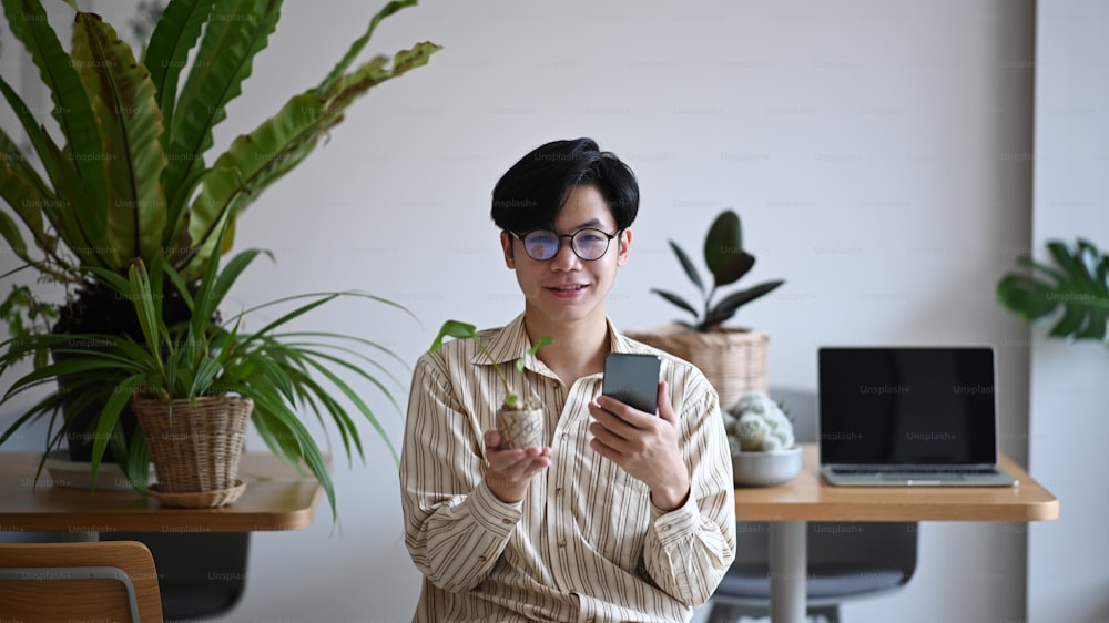 Smiling young man small business entrepreneur holding potted plant and smart phone.