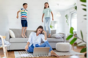Madre joven cansada sentada en el suelo y trabajando con computadora portátil y documentos mientras los niños pequeños saltan en el sofá y se divierten y hacen ruido