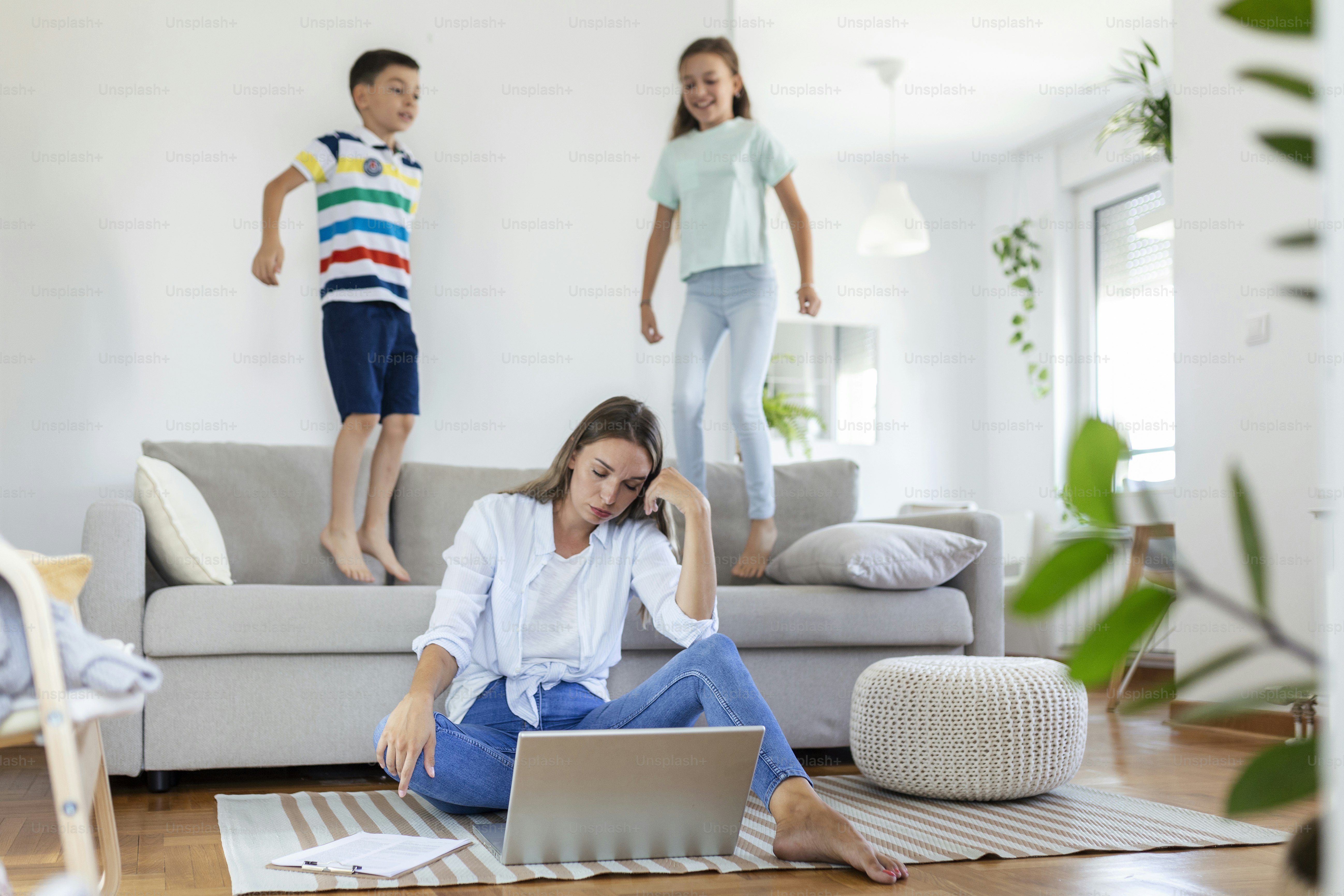 A woman using a laptop on the floor while two children jump on a sofa