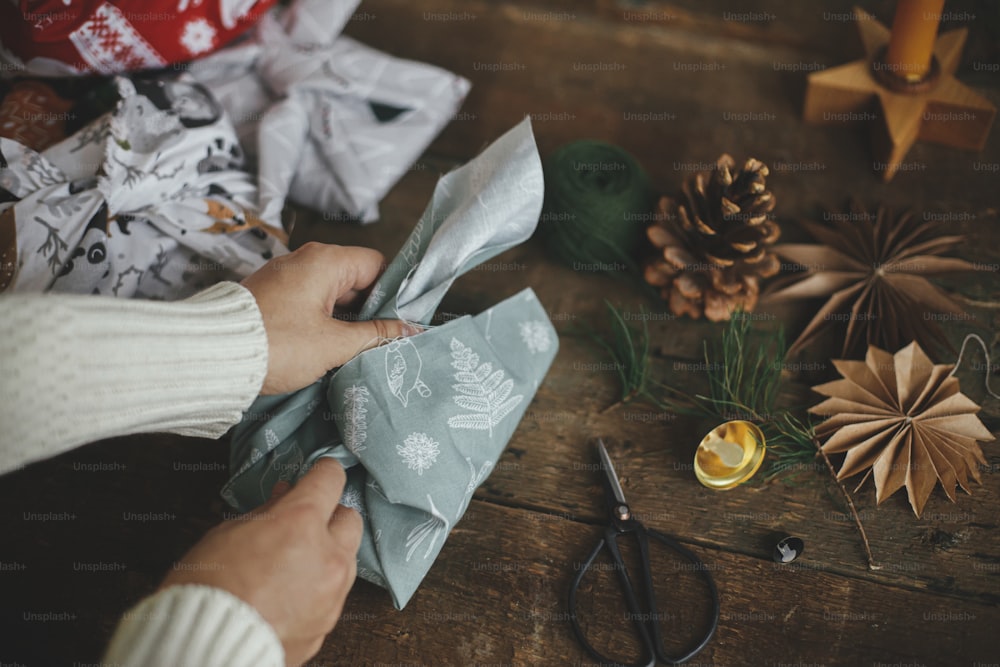 Weihnachtsgeschenk Furoshiki. Hände wickeln Weihnachtsgeschenk in Stoff auf rustikalem Holztisch mit Ornamenten. Atmosphärisches stimmungsvolles Bild, nordischer Stil. Null Abfall und umweltfreundliche Geschenke. Urlaubsvorbereitung