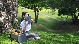 Peaceful young woman sitting under tree in the park and reading interesting book.