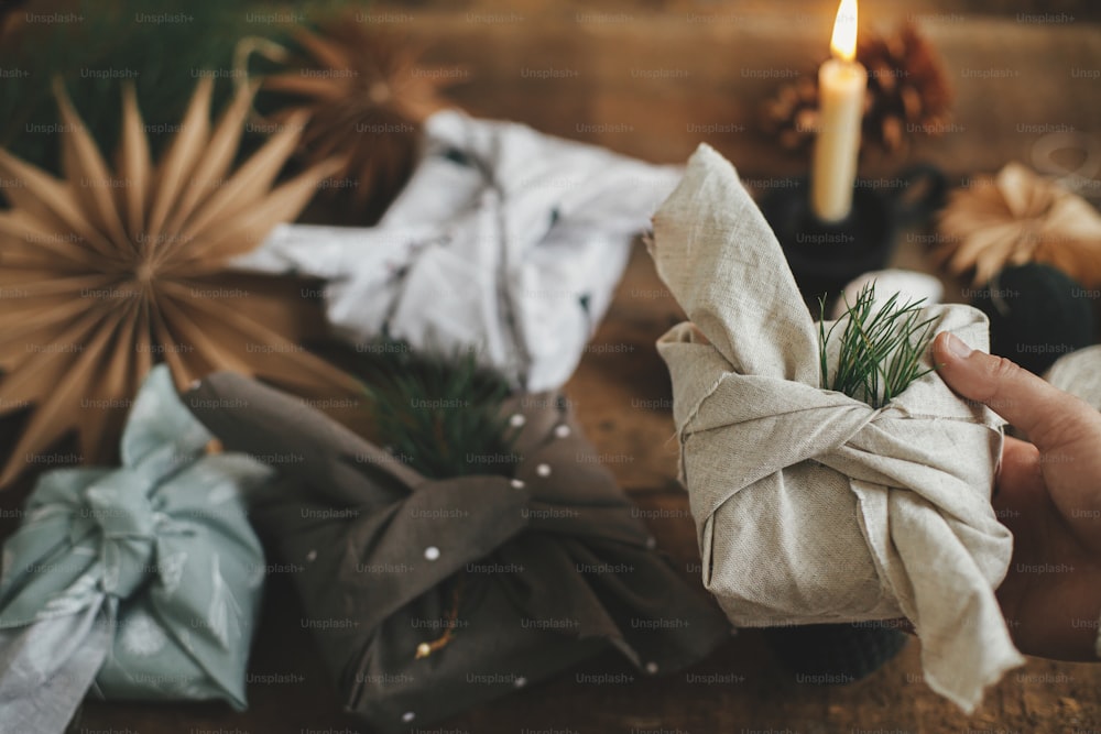風呂敷ラップ、ゼロウェイストのクリスマス休暇。オーナメント付きの素朴な木製のテーブルの上で、モダンなお祝いの生地に包まれたクリスマスプレゼントを持つ手。雰囲気のあるムーディーなイメージ、北欧スタイル
