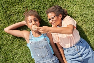 Kleines Mädchen, das Kaugummi ihrer Freundin im Freien zerbricht. Konzept der Kindheit. Idee der Freundschaft. Moderner Kinder-Lifestyle. Scherzhafte lockige brünette Mädchen, die auf grünem Gras liegen.