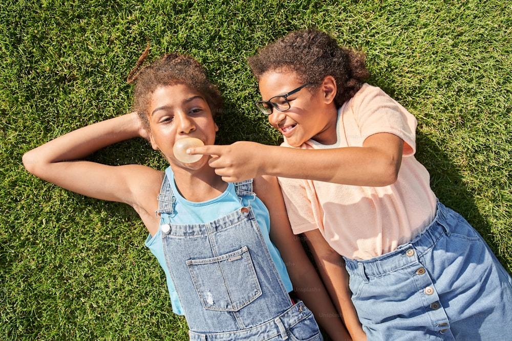 Little girl breaking chewing gum of her girlfriend outdoor. Concept of childhood. Idea of friendship. Modern kid lifestyle. Joking curly brunette girls lying on green grass.
