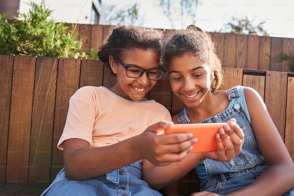 Kleine schwarze Freundinnen sitzen auf einer Holzbank und schauen sich draußen etwas auf dem Handy an. Konzept der Kindheit. Idee der Freundschaft. Moderner Kinder-Lifestyle. Lächelnde lockige brünette Mädchen. Sonnentag