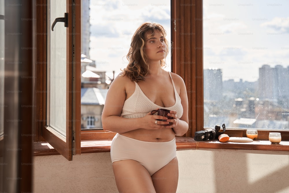 Oversize woman standing at opened window and drinking drink at home. Body positive. Idea of feminism. Domestic lifestyle. Young thoughtful european girl with tattoo. City view from window. Sunny day