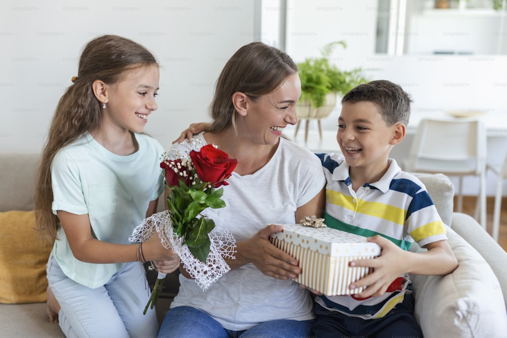 Fröhliches kleines Mädchen mit Geschenkbox und jüngster Bruder mit Rosenstrauß Blumen lächelnd und gratuliert gl�ücklicher Mama am Muttertag zu Hause. Alles Gute zum Muttertag!