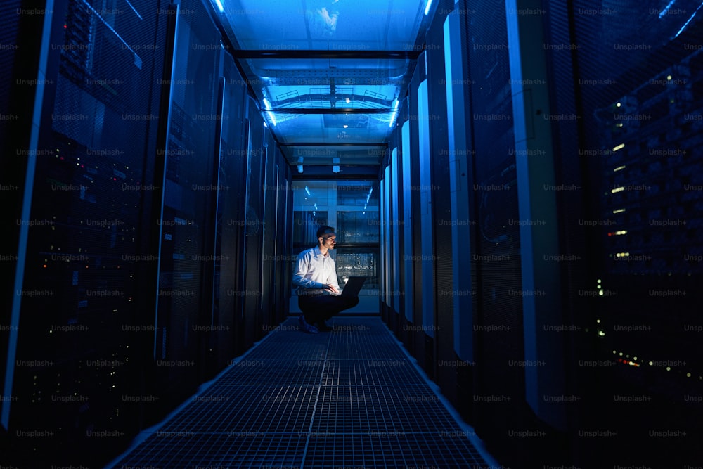 Data center administrator sitting on haunches before server rack and writing report on his laptop