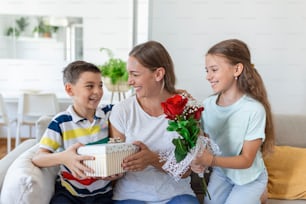 Happy children giving gift an flowers to mother. Happy Mothers Day! Children boy and girl congratulate smiling mother, give her flowers bouquet of roses and a gift box during holiday celebration