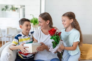 Glückliche Kinder, die der Mutter Blumen schenken. Alles Gute zum Muttertag! Kinder Junge und Mädchen gratulieren lächelnder Mutter, geben ihr Blumenstrauß Rosen und eine Geschenkbox während der Weihnachtsfeier