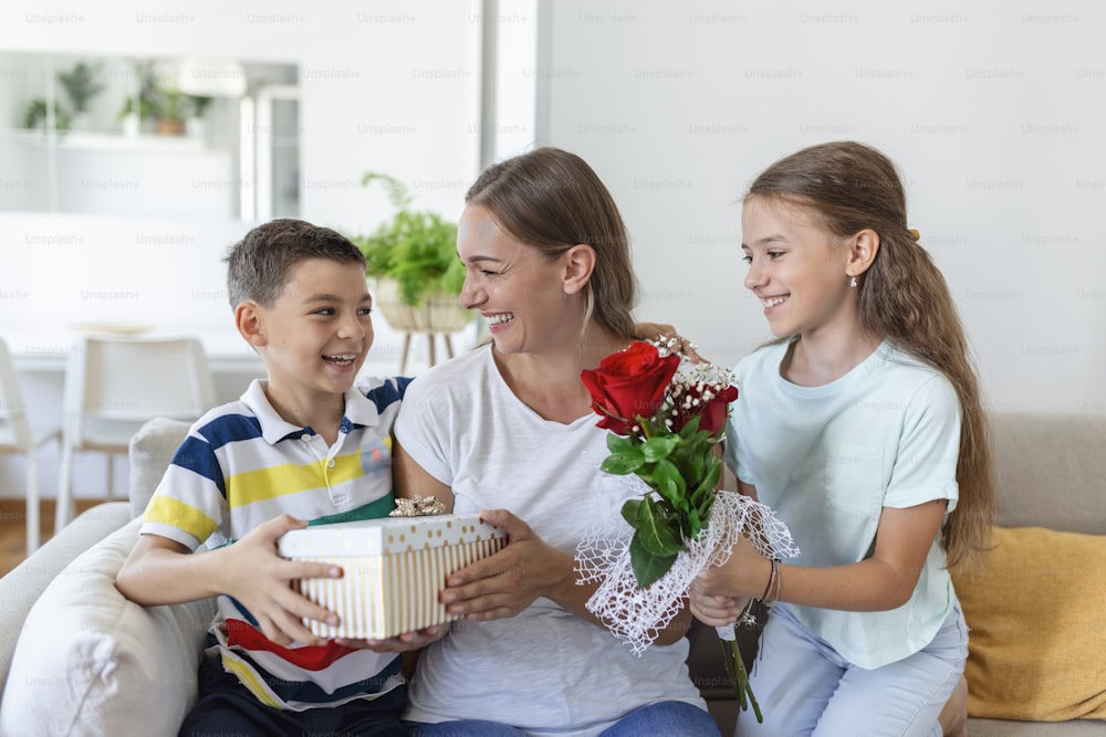 Happy children giving gift an flowers to mother. Happy Mothers Day! Children boy and girl congratulate smiling mother, give her flowers bouquet of roses and a gift box during holiday celebration