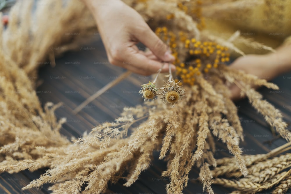 Stilvoller Herbst-Boho-Kranz mit trockenem Gras und Wildblumen auf rustikalem Holztisch. Ferienwerkstatt. Florist im gelben Pullover macht rustikalen Kranz auf dunklem Holz