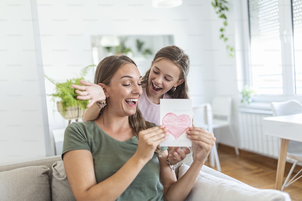 Young woman and girl at home celebrating mother's day sitting on sofa daughter hugging mother kissing cheek mom laughing joyful holding gift box