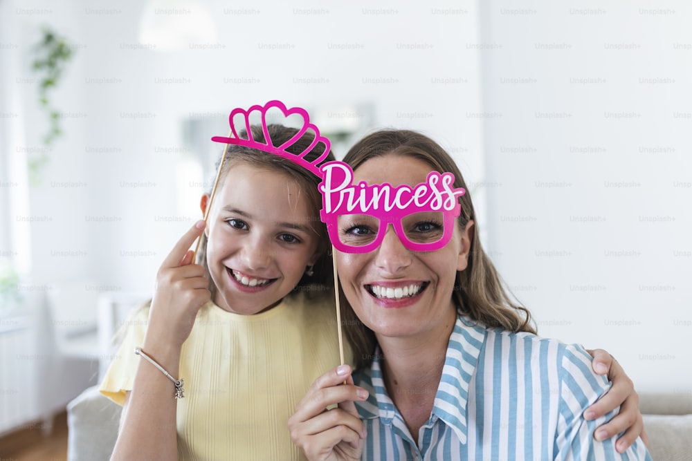 Funny family on a background of bright wall. Mother and her daughter girl with a paper accessories. Mom and child are holding paper crown on stick.