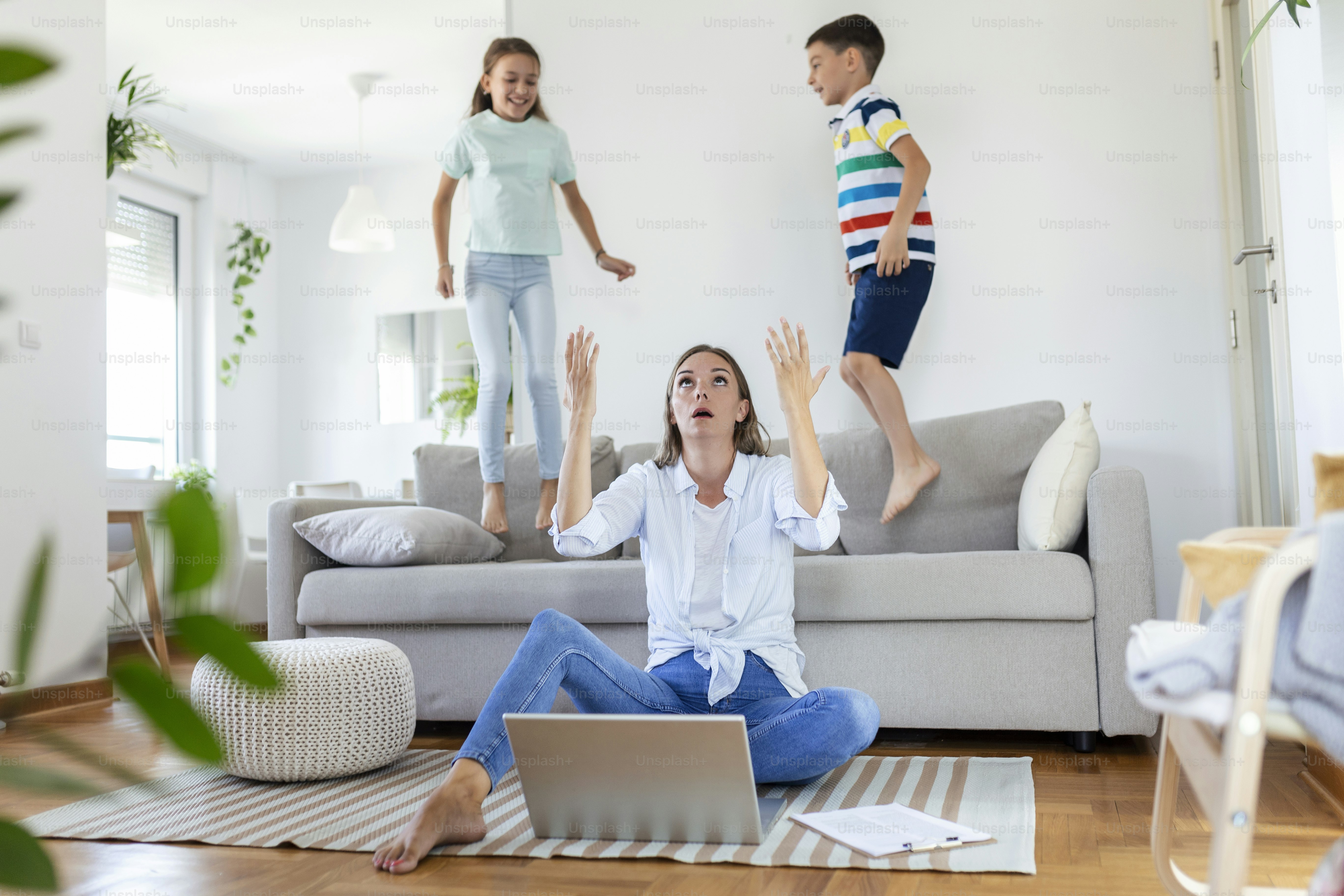 Tired young mother sitting on the floor and working with laptop and documents while little kids and jumping on the sofa and having fun and making noise